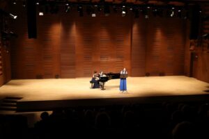 Na scenie śpiewaczka w niebieskiej sukni oraz pianistka za nią.