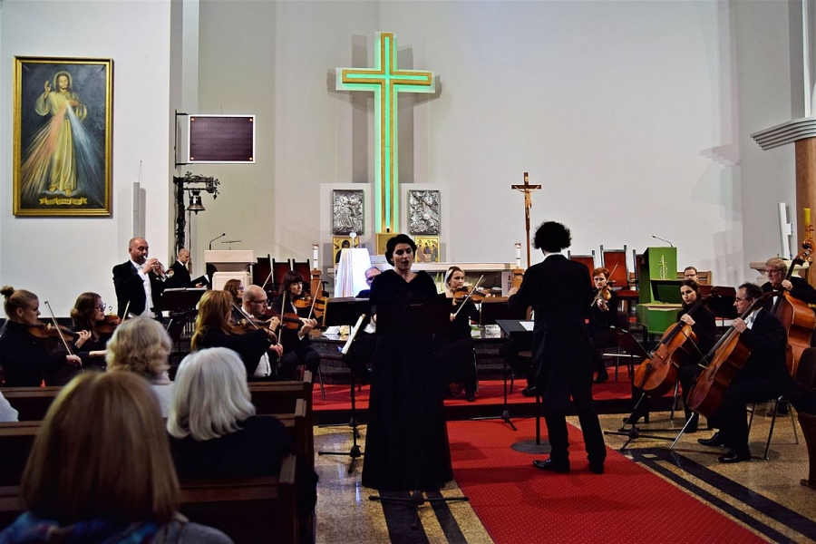 Na pierwszy planie sopranistka podczas występu, w tle orkiestra. Za nią pomnik krzyża podświetlony na zielono.